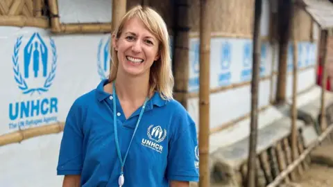 JOA Amber is wearing a blue UNHCR polo shirt stands in front of a row of bamboo-framed shelters covered with white tarpaulin displaying the UNHCR logo and text “The UN Refugee Agency.” The individual has a lanyard with an identification badge and is dressed in green trousers. The setting appears to be an outdoor area with a dirt pathway and trees visible in the background.
