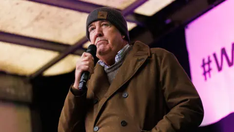 Dan Kitwood/Getty Images TV presenter Jeremy Clarkson, holding a microphone, speaks to the crowd at a farmers' demonstration in Whitehall, London in November 2024. He wears a brown coat and grey woolly hat.