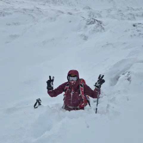 Catriona Burrow A woman wearing a dark pink jacket and black gloves standing in hip-deep snow. The snow is bright white. The woman has her hood up and goggles on. She is raising both her hands in an outwards facing peace symbol.