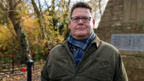 Kevin Laidlaw, a man in his 40s with short brown hair. He is wearing a smart green cord shirt and a blue tartan scarf. In the background is the war memorial in Swinton, with crocheted poppies tied to railings at intervals.