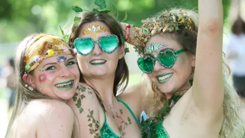 Jason Bryant Three young women pose for a photo at Shindig Festival. They are all wearing sparkly green outfits and have flowers and leaves painted on their bodies. 