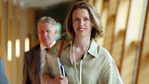 PA Media A woman in her 30s walking through a brightly lit corridor in the Scottish parliament. She is looking to the side and carrying a laptop and a notepad. The woman has brown hair and she's wearing a green shirt. There's a blurred man behind her.