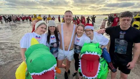 Jim Scott/BBC Un grupo de seis personas, dos de ellas con disfraces inflables de dinosaurio. El hombre del centro no lleva camisa. Todos sonríen a la cámara y se ve gente detrás de ellos en el mar.