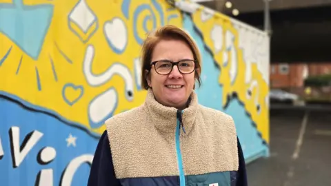 Paul Moseley/BBC Carli Harper is wearing a cream and blue fleece top. She is standing in front of a brightly decorated shipping container.