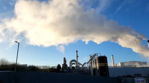 Ummer Daraz A factory plant with a large chimney spewing out white smoke on a clear day with blue skies.