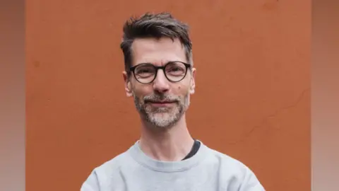 Tim Guy is standing in front of a bright orange wall. He is looking directly at  the camera and smiling. He is wearing a pair of round glasses and a grey pullover.