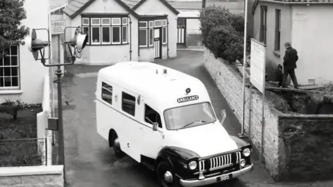 St John Ambulance and Rescue A black and white image of a very old fashioned white ambulance as it exits a driveway. There are houses on either side and a building with bay windows behind it.