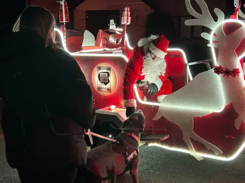 The sleigh from the above picture, lit up with bright white lights around the edge. Santa is sitting in the sleigh and leaning down towards a dog looking up at him from the ground.