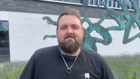 Luke is smiling at the camera, he has a dark navy t shirt on and a silver ring on a chain around his neck. He has short light brown hair and a moustache and beard. 