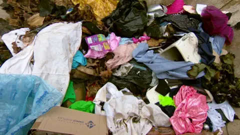 A skip is filled with discarded clothing and other rubbish. You can see various dance outfits in a variety of colours, including gold, pink, bright green and purple. There are also various bits what looks like scenery from dance productions, and cardboard boxes.