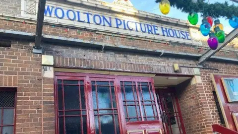 Art deco front of Woolton Picture House, which has its name sign in blue at the top of the single-stroey red brick building and red doors. Multi-coloured light bulbs hang from a green Christmas garland.