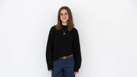Bath Spa University Students Union Mia Latkovic smiles at the camera as she stands in front of a white wall. She has shoulder-length brown hair and is wearing a black jumper and blue striped trousers.