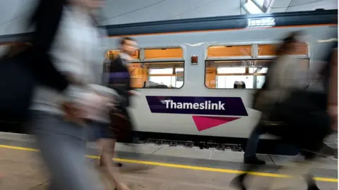 A white train with Thameslink written on its side. People are walking along the platform past the train, although they have been blurred.