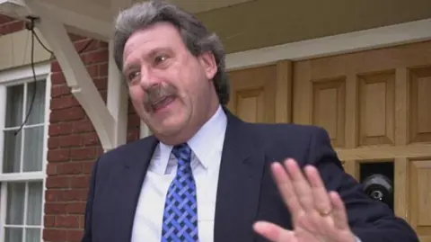 PA Media A man wearing a black suit jacket, white shirt and blue tie stands outside a house door. He is looking away from the camera with one hand up.