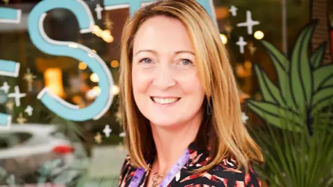 Aga Mortlock Sewing teacher Dawn Elliott with long blond hair wearing a black and orange patterned top with a lilac measuring tape around her neck. She is standing in front of a shop window with writing and green leaves painted on it. She is smiling. 