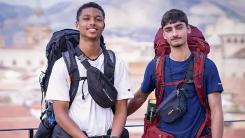 Jo and Kush, who are carrying large backpacks, are smiling at the camera at the start on the race in Italy. 