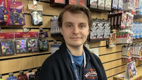 Darren Rozier/BBC A man stands in a cafe where trading cards and memorabilia have been placed on the wall behind him. He has dark hair and is smiling. He wears a black jumper.