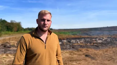 A man with blond hair stood in front of a burnt field. He's wearing a mustard yellow quarter-zip. The sky above is bright blue. 