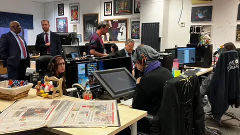 An office with five people sitting at computers, while two men in suits stand talking to the left
