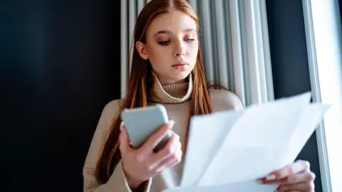 Getty Images Young woman with red hair, wearing beige jumper holds smartphone in one hand and household bills in the other 