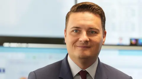 Wes Streeting is smiling and dressed in a navy suit with a light blue shirt and maroon tie. His short brown hair is neatly combed back from his face.