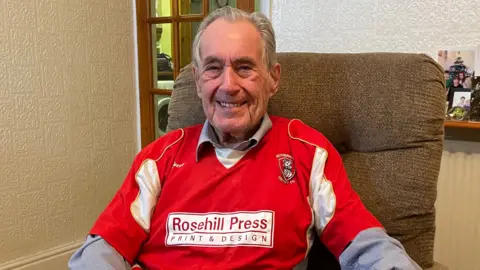 Tom MacDougall / BBC Dennis, an elderly man, smiling and wearing a Rotherham United shirt.