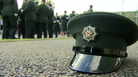 A PSNI hat sits on the ground n the foreground of the picture while male and female new police officers stand in the background in their uniforms