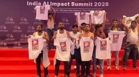 Eleven men on a red carpet in the AI Summit venue in Delhi on 20 February. At least seven of them are shirtless. Nine of them are holding tee shirts with printed images of PM Narendra Modi and US President Donald Trump.