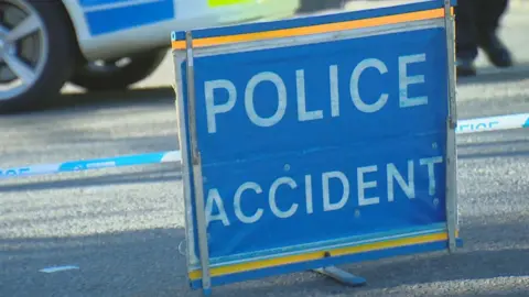 A blue police sign that reads 'Police Accident' on a road. In the background there is a parked police car and police tape.