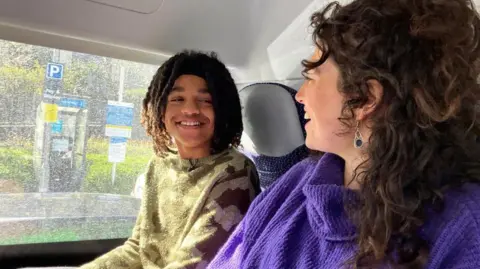 A teenage boy in a green jumper sitting next to a woman on a bus who is wearing a purple jumper. They are smiling at each other. The bus window is dirty. In the background, outside of the bus, you can see signs and greenery. 