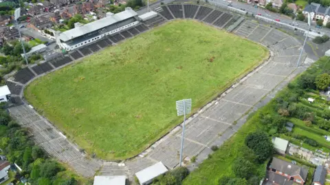 PA Media An aerial view of a grass pitch with concrete terrace-style stands. It sits in a residential area with a number of red brick houses surrounding the site. 