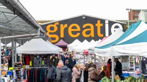 Customers browse market stalls selling clothing, fruit and electrical goods, with a large sign reading 'greatie' in orange and white lettering in the backdrop.