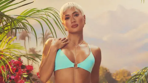 ITV Kaz Crossley in a promo shot for Love Island All Stars. Kaz has a blonde pixie cut and wears a light blue bikini, her right hand resting on her right shoulder. She's surrounded by tropical plants. 