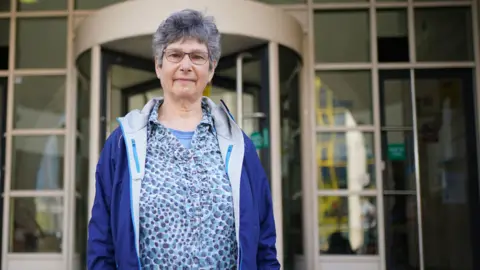 A woman in her 60s with grey hair and wearing a blue waterproof jacket and a blue, purple and white patterned top. She is standing outside the entrance to a building with the blurred image of a metal and glass revolving door and windows behind her.