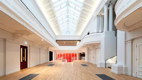 Renfrewshire Council Inside one of the new display galleries at the Paisley Museum looking back towards the red glass entrance. The walls are painted white, there is a wooden floor and sky lights on the ceiling.