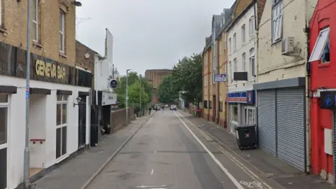 Google Geneva Street in Peterborough, showing Geneva Bar to the left, with a building to the right with metal shutters across its windows and a large black wheelie bin outside. A takeaway shop is next door. The single lane road is empty, with no traffic or people.