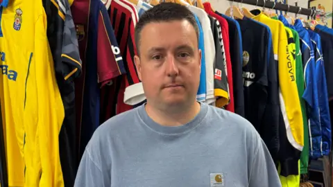 A man with dark hair wearing a blue top looking at the camera