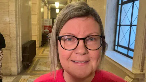 A blonde woman wearing glasses, she is smiling at the camera and wearing a red top. 