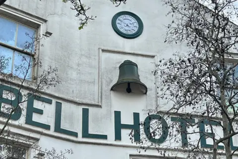 A circular clock above the Bell Hotel sign on the side of the cream building displays the time as 13:50.