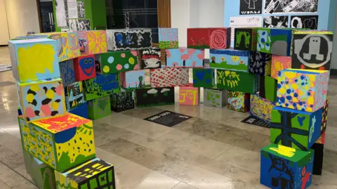 Colourful square boxes that have been painted on by school children. They are all stacked together in a circle shape.