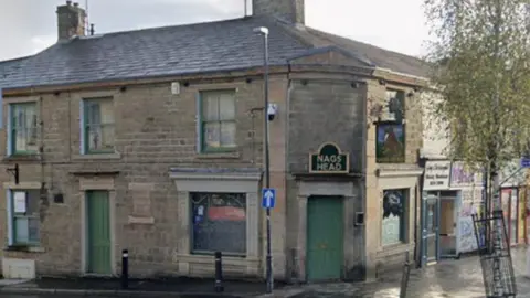 Street view image of the Nag's Head pub. A stone building on a street corner with green windows and doors and a sign in black with gold wording over the door, which is on the corner, that says NAG'S HEAD