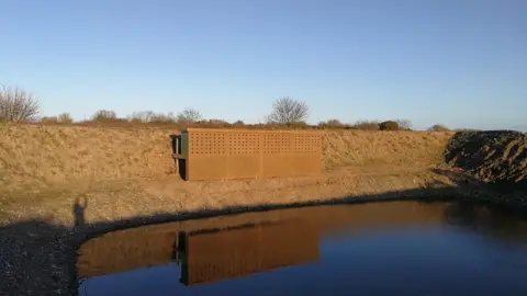 The wooden structure which is attached to a bank, it camouflages in with the surroundings and has small holes, there is a body of water in front on a clear blue day.