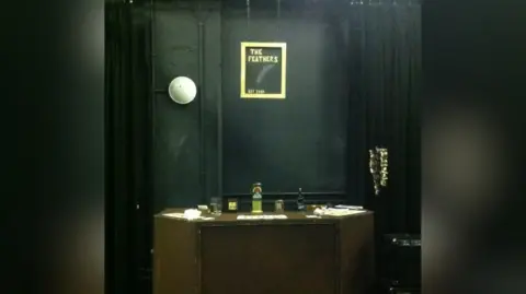 Roisin Quinn A theatre set bar with a sign reading 'The Feathers' in a gold frame. The walls are painted in a dark charcoal colour and there is a wooden bar with beer pumps. 
