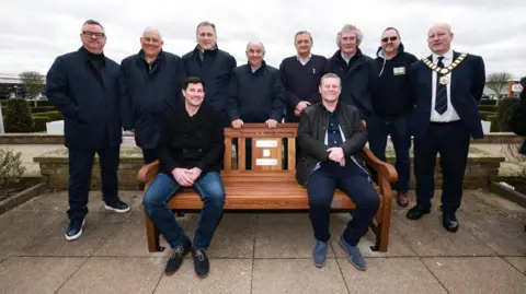 Several people gather around a brown wooden bench. Eight people are stood behind it, with two people sat on the bench. On the bench there are several plaques, and above them is some black writing which says "someone is always listening".