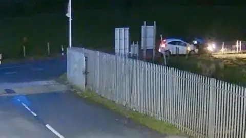 A road with a metal fence which is covering a white car with its lights on 
