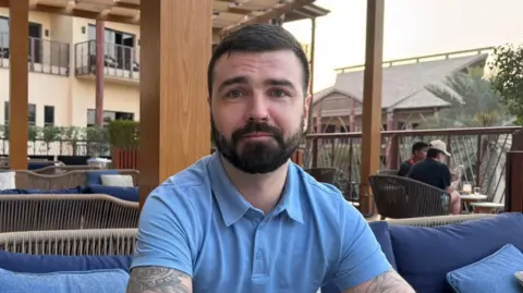 Mic Cassidy has dark hair and a dark beard, tattoos on his arms and is wearing a blue, short-sleeved top, sitting in a hotel cafe area