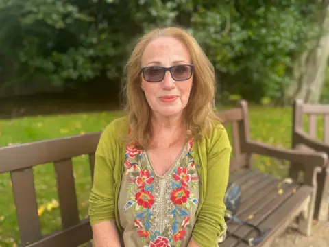 May Hooper wears a lime greenish  cardigan and floral top, sitting connected  a seat  successful  a park