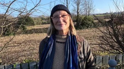 Barbara has blonde hair down to her shoulders and wears glasses, a black beanie hat, blue scarf, brown jacket and grey jumper. She is smiling while she stands in front of the fields.
