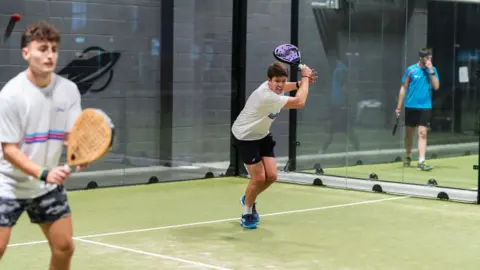 UK Padel Three men holding sports rackets in a padel court. The playing surface is green with white lines