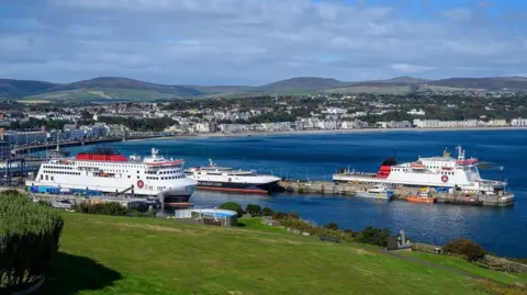 Three ferries, with red white and blue livery, moored in Douglas Harbour. There is green grass in the foreground and the sea is blue. In the background, lots of houses and building line the sea front, with rolling hills behind them.